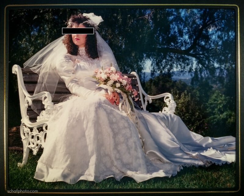 A bride in a white wedding gown and veil sits on a white ornate bench outdoors, holding a bouquet of pink and white flowers, with greenery and trees in the background. Her eyes are concealed for privacy.