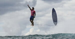 A surfer in a pink shirt and blue shorts appears to be floating above the ocean, raising one arm, while his surfboard is airborne nearby, attached by a leash. The sky is cloudy.