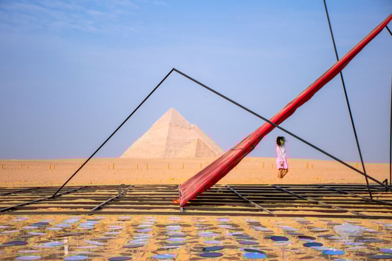 Una joven camina frente a una instalación de arte con espejos y tela roja en el desierto cerca de la Gran Pirámide de Giza bajo un cielo azul claro.
