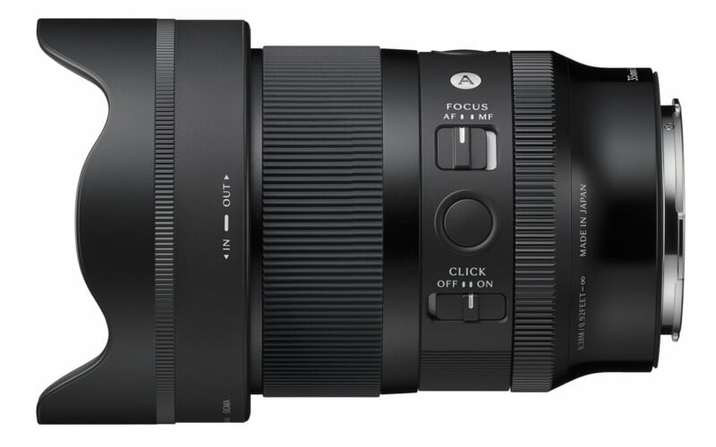 A close-up side view of a black camera lens with a lens hood attached, showing focus and click switches, a focus mode dial, and detailed markings on the lens barrel.