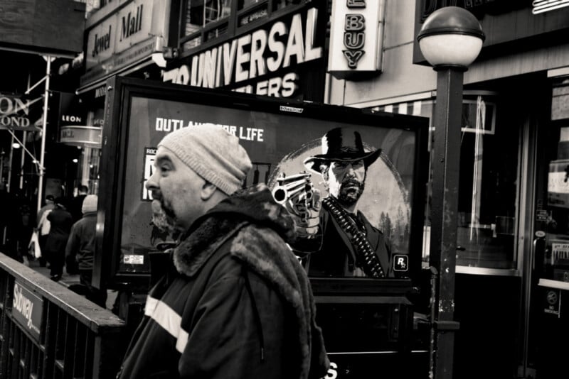 Un hombre vestido con un abrigo de invierno y un sombrero caminaba por la entrada del metro, detrás de él un gran cartel que mostraba a un vaquero apuntando con un arma. La escena está llena de escaparates y gente al fondo.