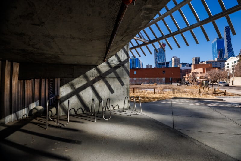 A sunny urban scene shows empty bike racks under a concrete overhang, casting shadows on the ground. In the background, modern city buildings and a blue sky are visible beyond an open, grassy lot.