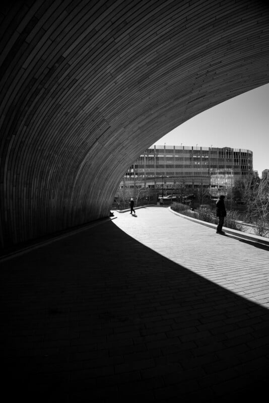 A curved wooden structure casts a dramatic shadow on a walkway, dividing the scene into light and dark. Two people stand in the distance, with a modern building and trees visible under a clear sky.