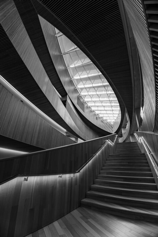 A monochrome architectural photo of a modern, curved staircase with wooden paneling and a geometric skylight, creating dramatic shadows and sweeping lines.
