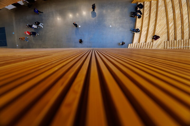 A view from above shows people walking on a gray floor beside wide wooden stairs. Vertical wooden slats in the foreground create a strong geometric pattern leading toward the scene below.
