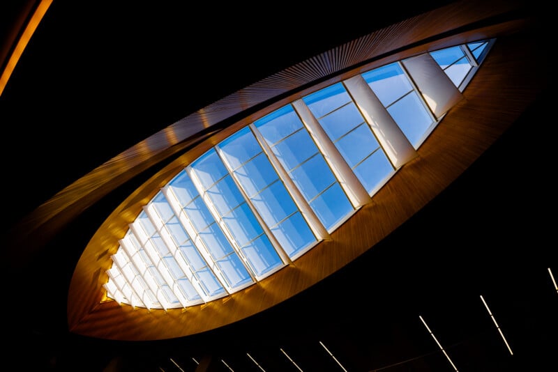 A large elliptical skylight with a wooden frame lets natural light through angled glass panels, showing a blue sky. The skylight contrasts with the dark ceiling around it.