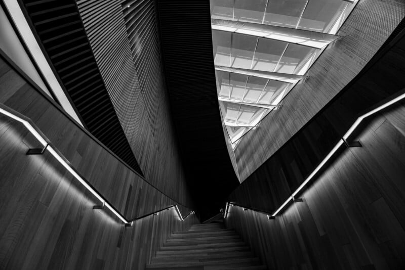 Black and white photo looking up a modern, angular staircase with wooden walls and illuminated handrails, ending at a ceiling with geometric skylights and strong architectural lines.