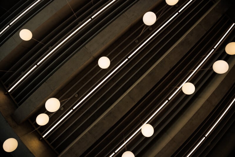 Round, glowing light fixtures hang from the ceiling in diagonal rows. The ceiling consists of gray, parallel beams, creating a geometric, modern architectural pattern. The lights appear as bright orbs.