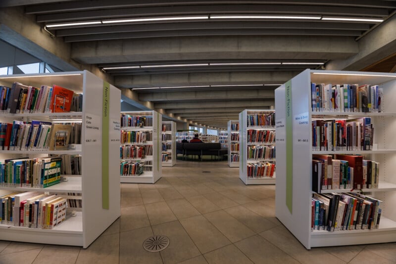 Wide-angle view of a modern library interior with rows of white bookshelves filled with books, tiled floor, and overhead concrete beams. In the background, a person sits reading on curved chairs.