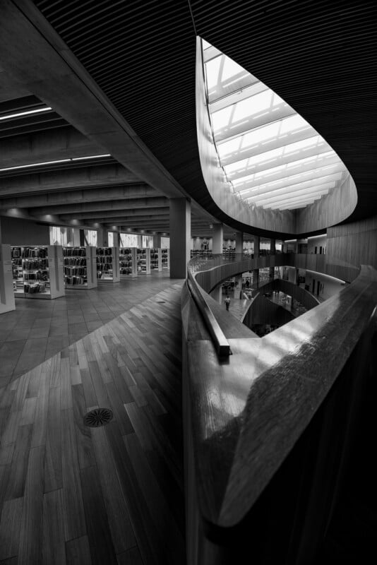 Black and white photo of a modern library interior with curved wooden balcony, bookshelves, skylights, and sleek architectural lines creating dramatic contrasts and shadows.