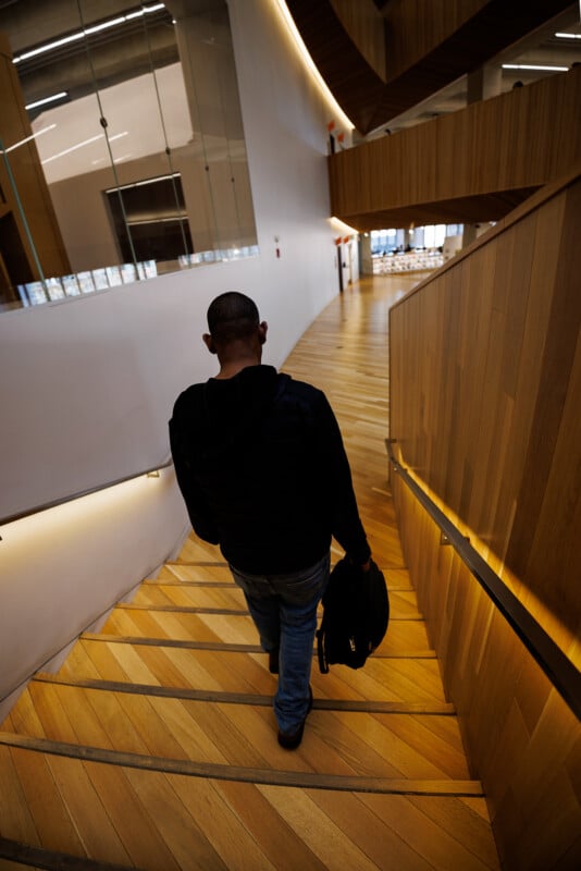 A person wearing a dark jacket and jeans walks down wide wooden stairs in a modern building, holding a black bag; the space features curved white walls and a sleek, open design.