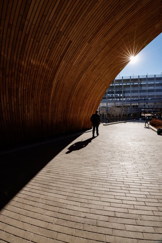 A person stands under a large, curved wooden structure casting a long shadow on the brick pavement. Sunlight shines through, and a modern building is visible in the background.