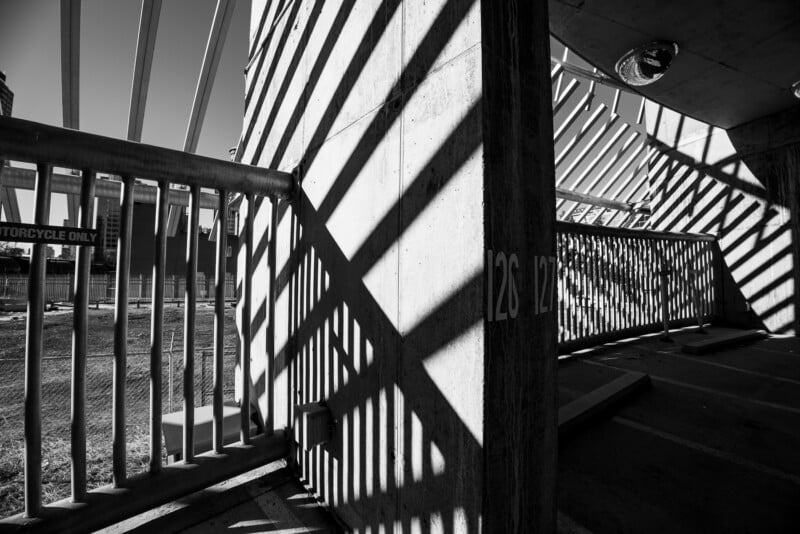 Black and white photo of strong shadows from railings creating diagonal patterns on a concrete pillar and ground in a parking garage, with sunlight streaming through. "Motorcycle Only" sign visible outside.