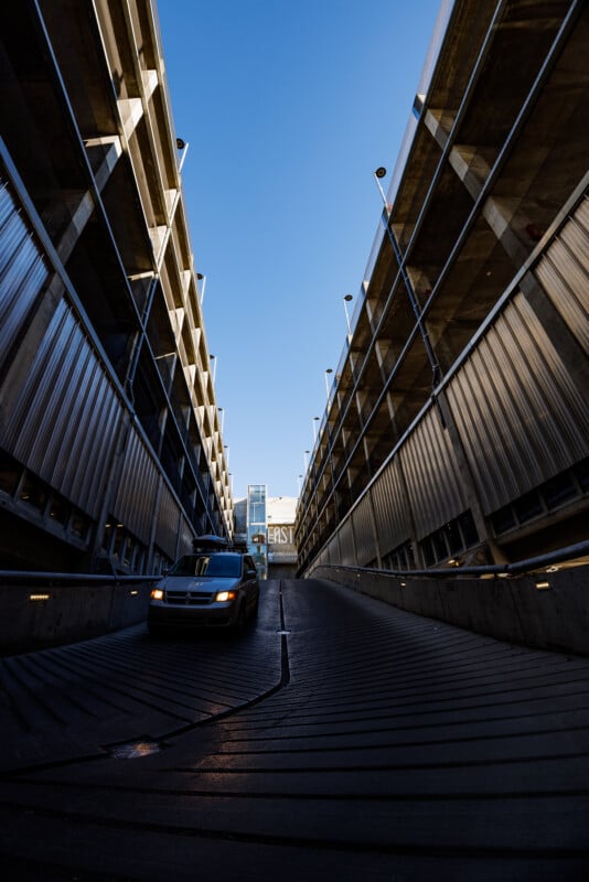 A black car drives up a sloped ramp between two concrete parking garage structures under a clear blue sky, with a glass building visible in the background.