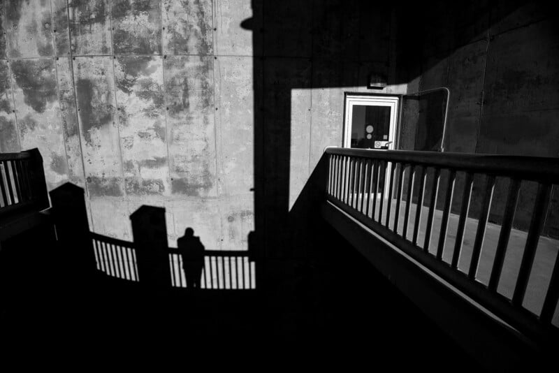 A shadow of a person is cast on a concrete wall and railing in a stark, high-contrast outdoor scene, with bright sunlight creating sharp geometric shadows and a closed door in the background.
