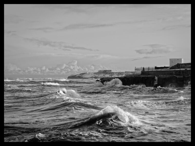 Black and white photo of rough waves crashing against a rocky shoreline, with some buildings and a distant cliff in the background under a cloudy sky.