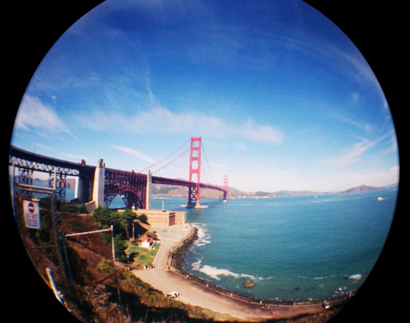 A fisheye lens photo of the Golden Gate Bridge spans over blue water and connects green hills under a bright, partly cloudy sky in San Francisco. A road and shoreline are visible in the foreground.