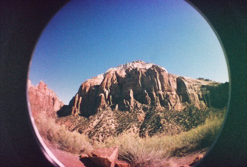 A rugged, rocky mountain under a clear blue sky, viewed through a circular fisheye lens with some brush and rocks in the foreground. The image has a retro, grainy effect.