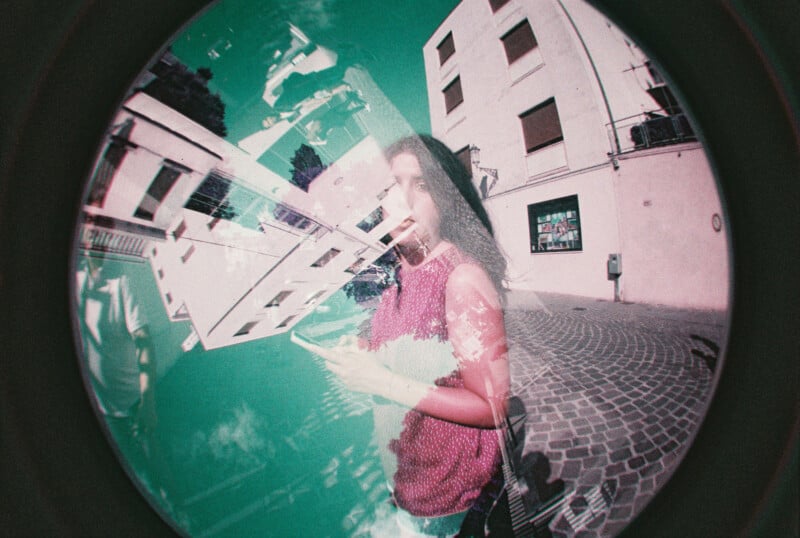 A fisheye lens photo with a double exposure effect shows a woman in a pink top holding a phone on a cobblestone street, overlaid with geometric shapes and building fragments, creating a surreal, dreamlike atmosphere.