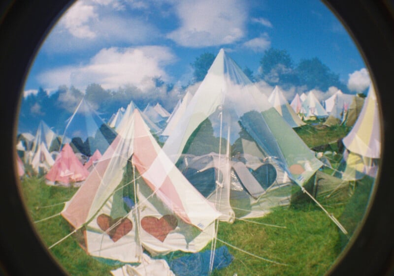 A fisheye lens photo shows colorful triangular tents with heart-shaped windows on a grassy field under a blue sky with clouds. Some trees are visible in the background, creating a whimsical, dreamy atmosphere.