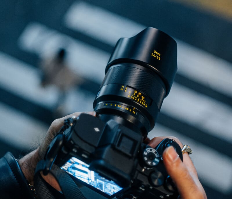 A person holds a digital camera with a large Zeiss Otus 1.4/35 lens, preparing to take a photo on a street with blurred white crosswalk lines in the background.
