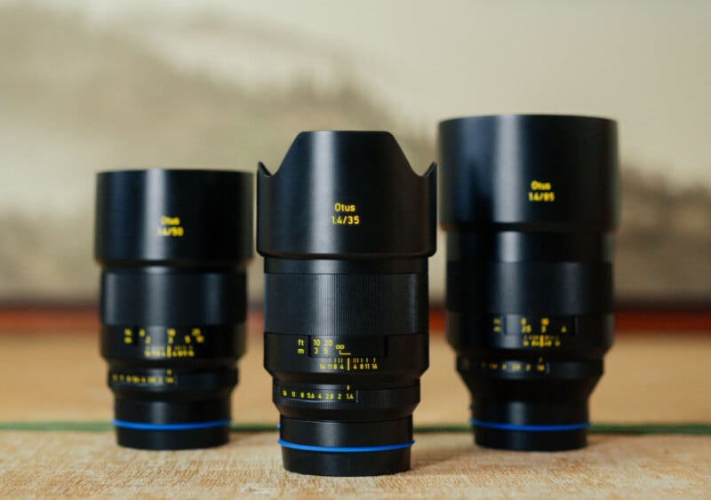 Three black Zeiss Otus camera lenses with yellow lettering are arranged side by side on a wooden surface, with the middle lens slightly taller and featuring a lens hood. The background is softly blurred.
