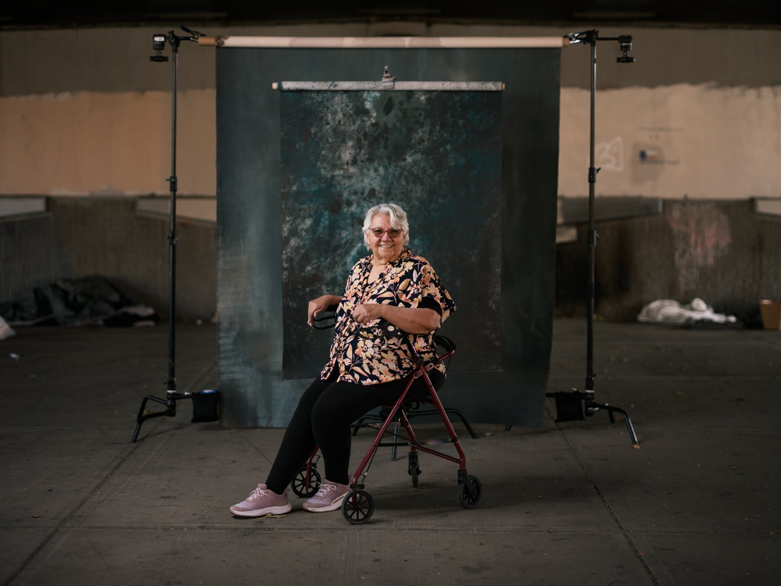 An older woman with gray hair and glasses sits smiling on a walker in front of a dark textured backdrop, set up in an urban outdoor space. She wears a floral shirt, black pants, and pink shoes.