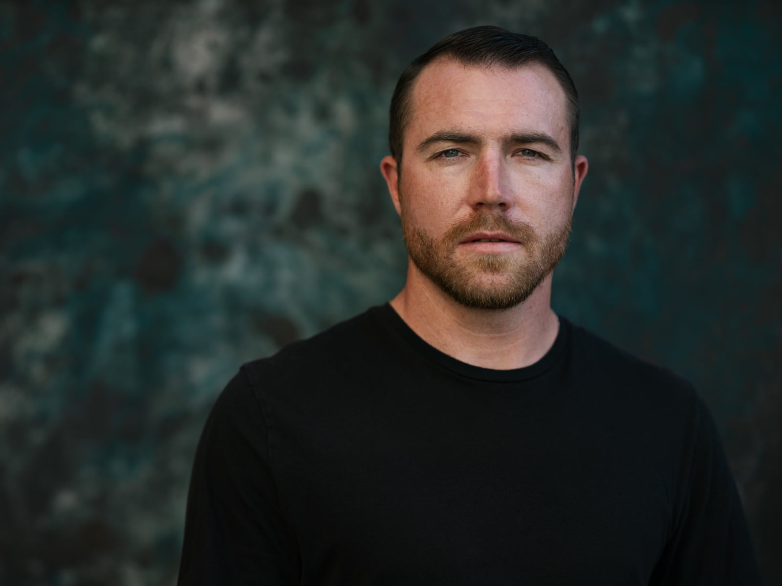 A man with short brown hair, a trimmed beard, and a serious expression is wearing a black shirt, standing in front of a blurred dark, textured background.