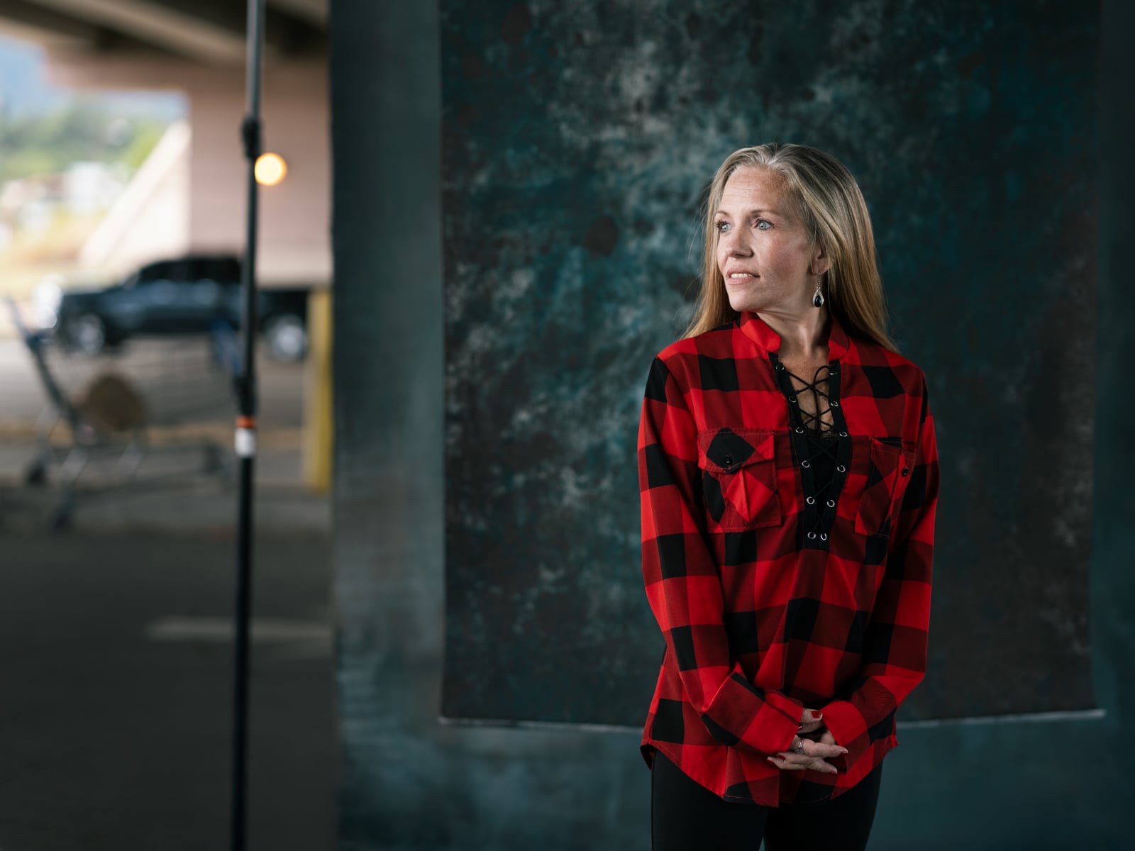A woman with long blond hair wearing a red and black plaid shirt stands with her hands clasped, looking to the side. She is under an overpass with blurred urban elements in the background.