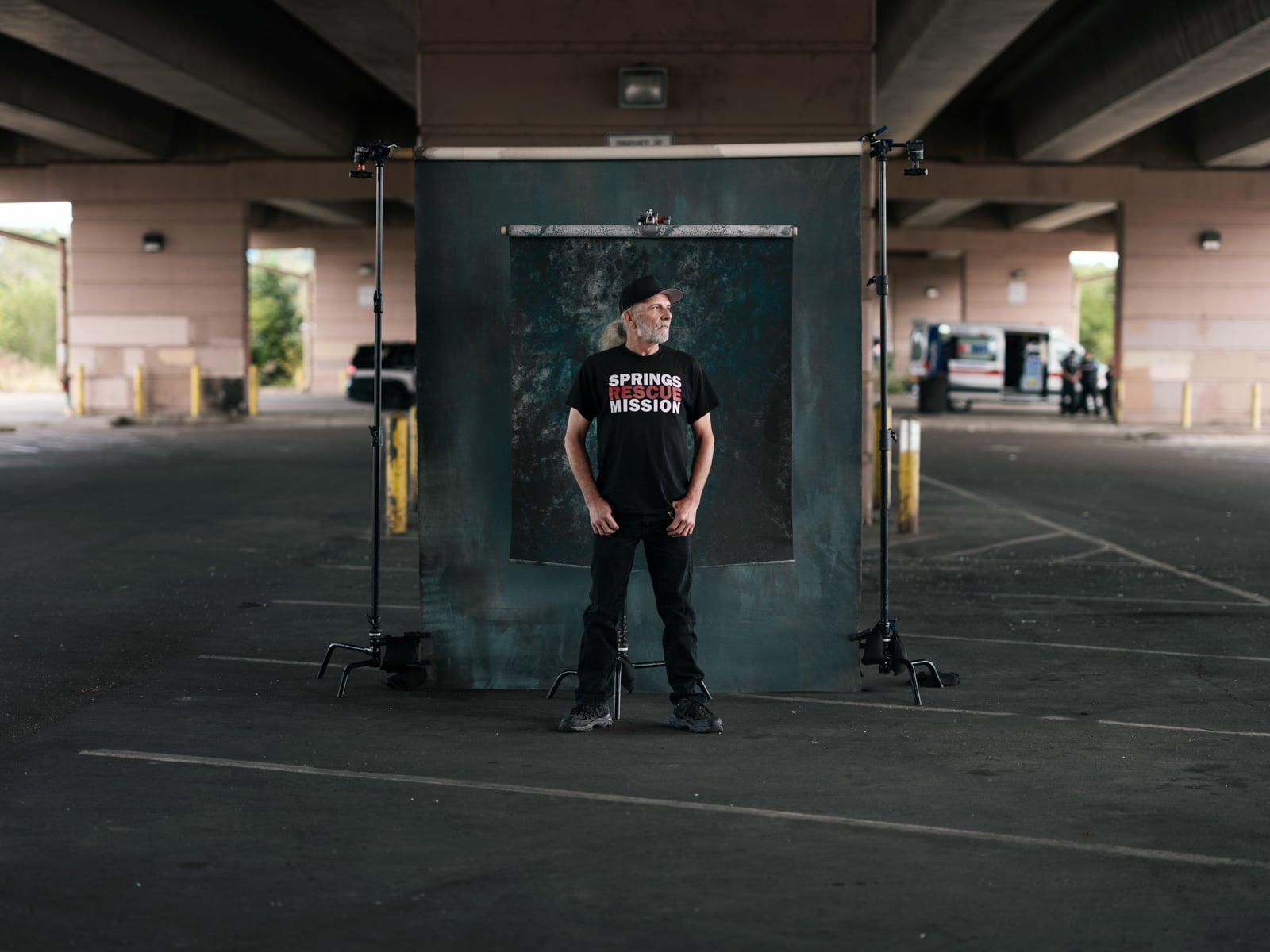 A man in a "Spring Mission" t-shirt stands in front of a backdrop under a highway overpass, with light stands on either side. An ambulance is visible in the background near the edge of the parking lot.