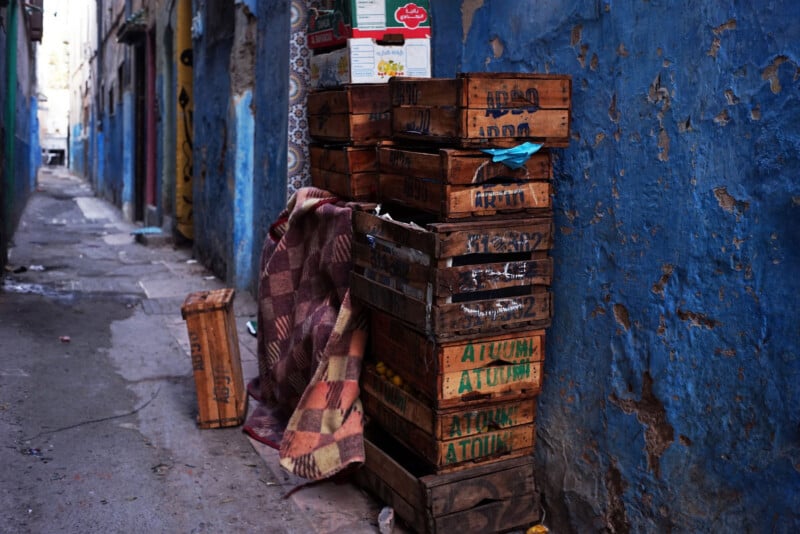 A stack of wooden crates and a closed box stand beside a worn blue wall in a narrow alley. A plaid blanket partially covers the crates, with scattered debris on the ground nearby.