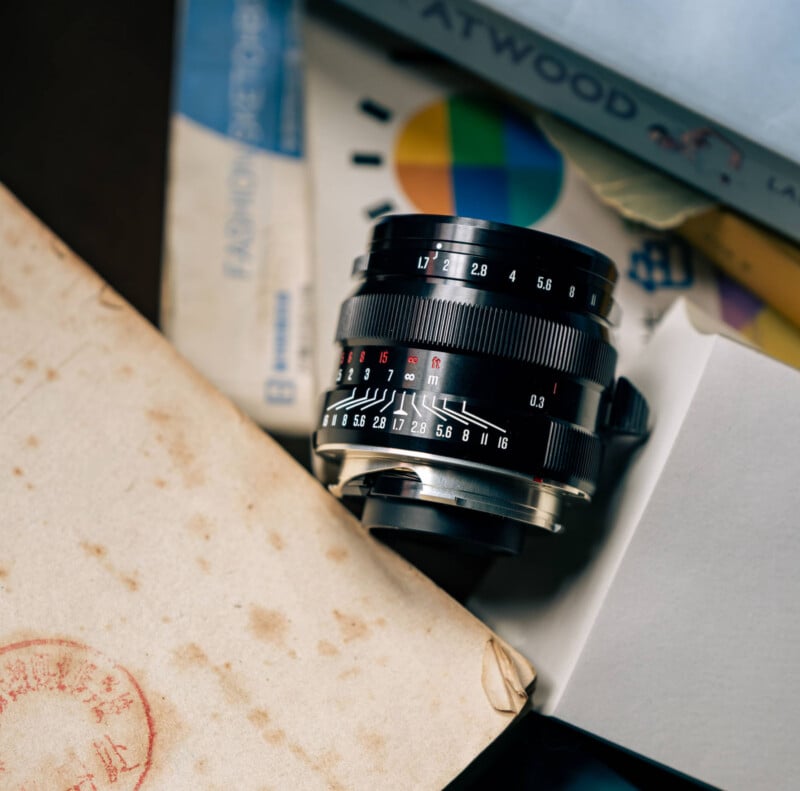 A close-up of a camera lens with visible aperture and focus markings, partially surrounded by old envelopes, books, and packaging. The lens is black with white and red numbers and sits at an angle among the clutter.