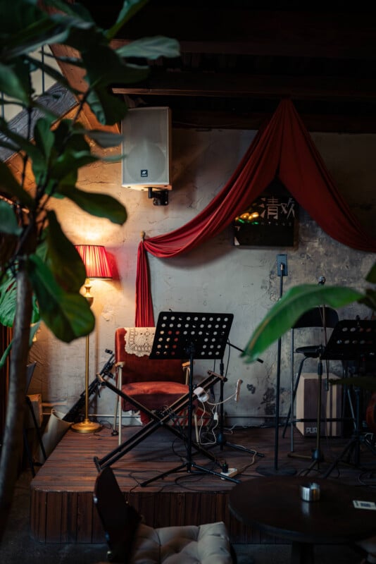 A small, cozy stage with a red armchair, music stands, a floor lamp, and red drapery against a textured wall, surrounded by potted plants in a warmly lit indoor setting.