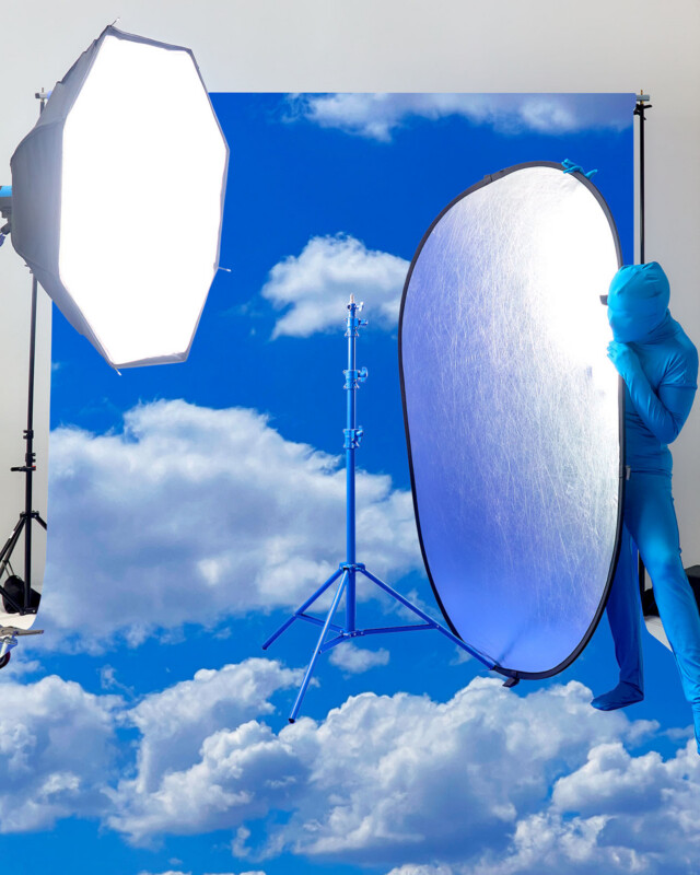 A person in a blue bodysuit holds a reflective panel in front of a cloudy sky backdrop, with a large studio light and stand also visible in the photography setup.