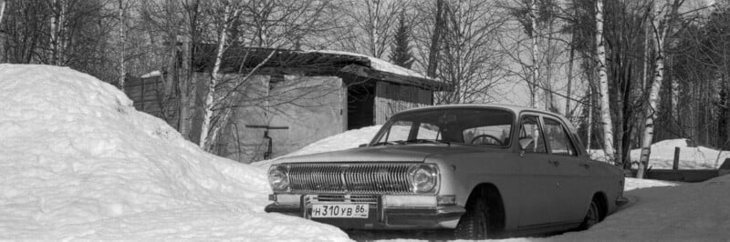 A vintage car is parked in deep snow near a wooden shed, surrounded by leafless trees on a bright winter day.