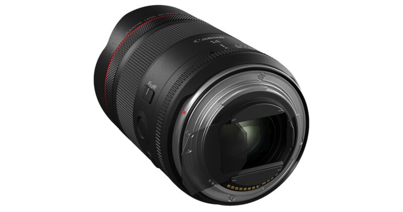 A close-up view of a Canon camera lens, showing the lens mount, glass elements, and textured focus ring against a white background.