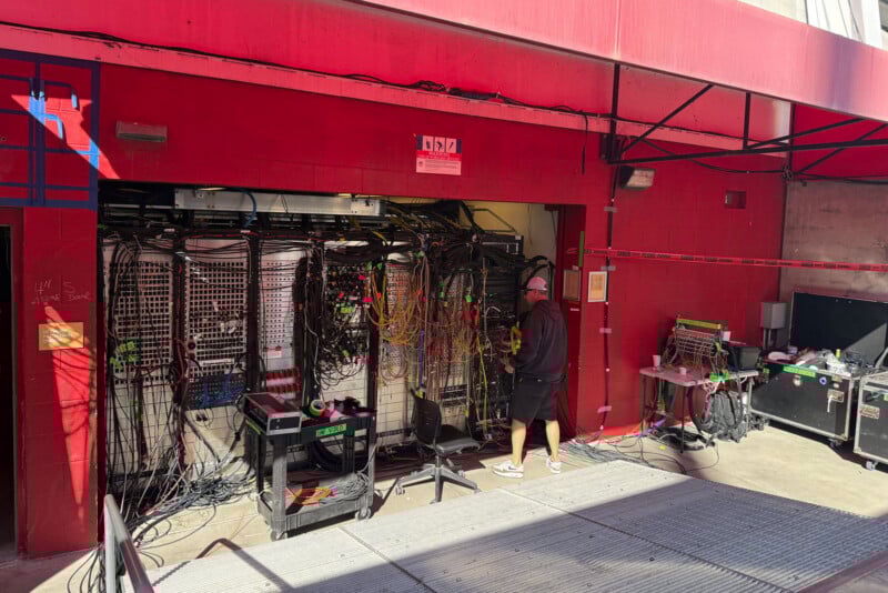A person wearing black works in front of a large setup of cables and electronic equipment inside a red-walled garage or booth, with sunlight casting shadows on the ground outside.