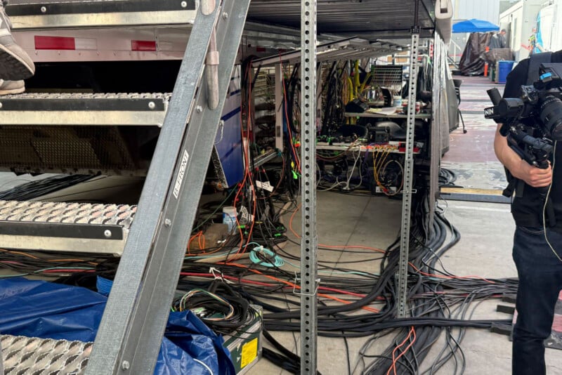 A behind-the-scenes view of a film or TV production setup, showing metal stairs, racks filled with tangled cables, electronic equipment, and a person holding a camera on the right edge of the image.