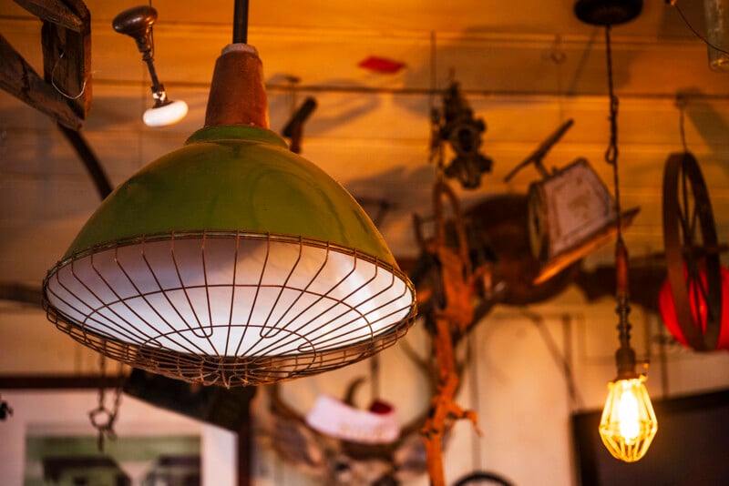A green industrial-style hanging lamp with a wire cage is lit in the foreground, while a warm exposed bulb and various vintage objects are blurred in the background.