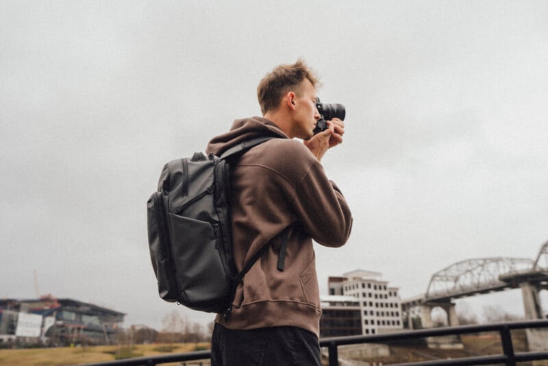 A person wearing a brown hoodie and black backpack is taking a photo with a camera outdoors, with a bridge and buildings visible in the background under a cloudy sky.
