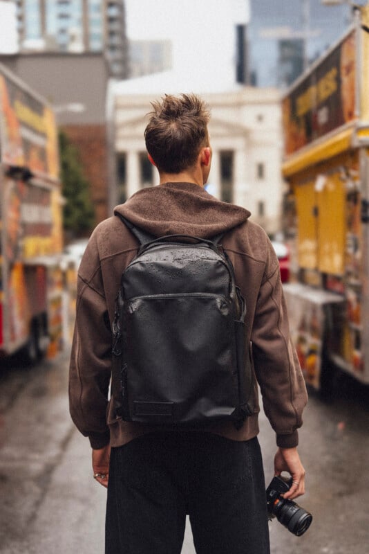 A person with short hair, wearing a brown hoodie and black backpack, stands in a city alley between food trucks, holding a camera in one hand. The ground appears wet, and buildings are visible in the background.