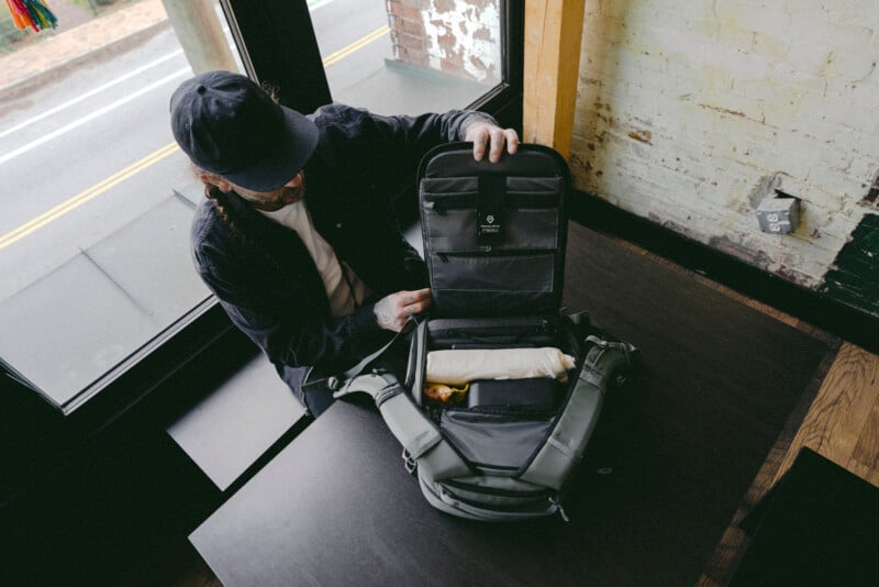 A person wearing a cap and glasses sits at a table by a window, opening a green backpack to reveal neatly packed items inside, including a rolled-up white cloth and a yellow pouch.