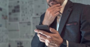 A man in a suit holds a smartphone in one hand and touches his chin thoughtfully with the other, standing in front of a wall covered in newspaper pages.