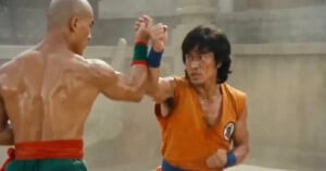 Two men are engaged in a martial arts fight, wearing sleeveless outfits with colored belts and wristbands. One has short hair and is shirtless, the other has shaggy hair and an orange shirt with a round emblem.