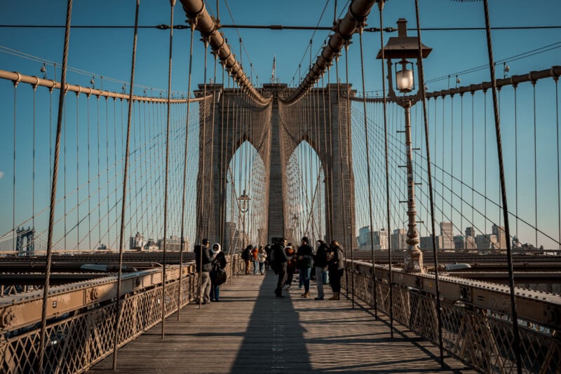 People walk along the pedestrian path of the Brooklyn Bridge in New York City on a clear day, with the bridge’s iconic stone arches and cables prominently visible. City buildings can be seen in the background.