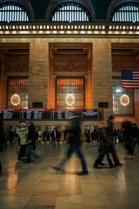 Commuters walk through the grand hall of a train station with high windows, chandeliers, an American flag, and electronic schedule boards. The lighting is warm and there is a sense of motion among the people.