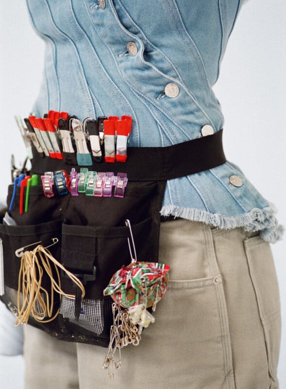 A person wearing a denim jacket and beige pants has a black utility belt around their waist, holding colorful clothespins, rubber bands, pens, and a bundle of wires.