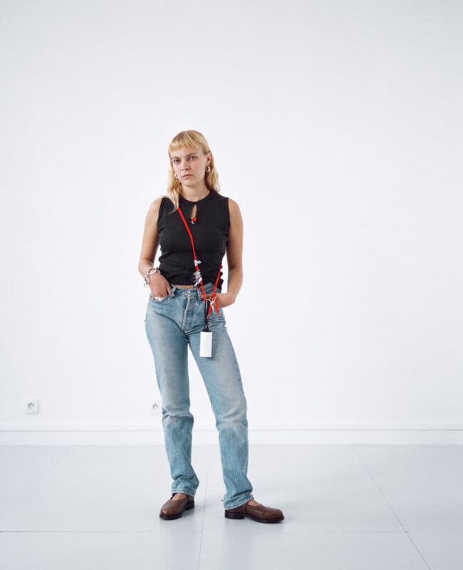 A person with light hair, wearing a black sleeveless top, light blue jeans, brown shoes, and a red lanyard stands in a minimalistic, white room with hands in their pockets, looking slightly to the side.