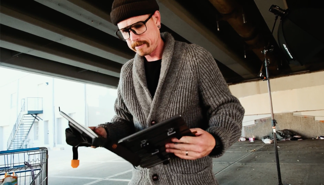 A man wearing glasses, a beanie, and a gray cardigan stands under an overpass, looking at a device in his hands with a focused expression.