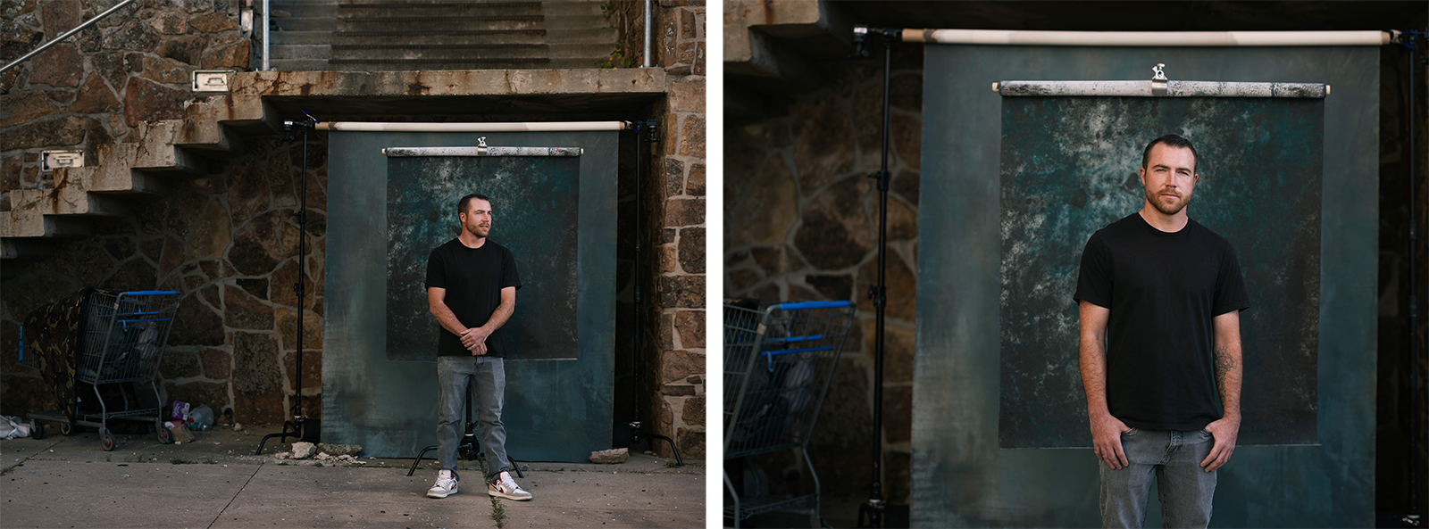 A man in a black t-shirt and jeans stands in front of a textured backdrop set up outdoors beside stone steps and a wall, with a blue shopping cart and debris visible nearby.